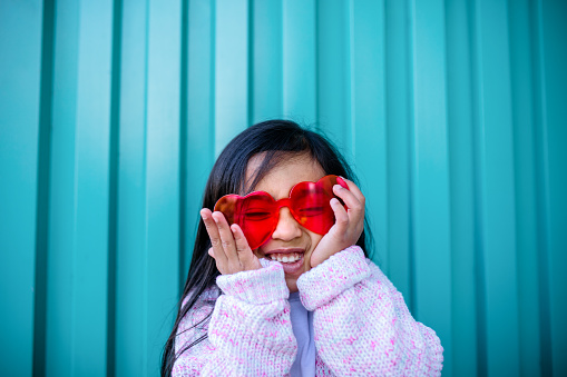 A cute mixed ethnicity child wears a pink sweater and glasses shaped like hearts for Valentines day.  Teal colored background.