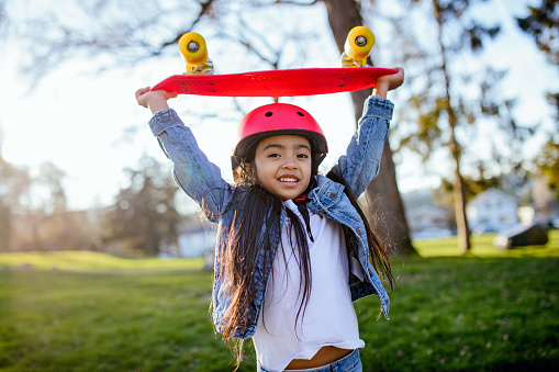 A cute elementary aged girl plays outside, enjoying skateboarding on a sunny day.  She wears blue denim and a red helmet that matches her skateboard.