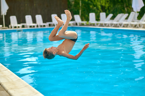 Flip jump. School-age boy in air upside down above water of outdoor pool at moment of flip jump