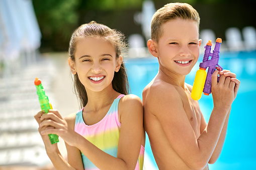 Summer. Smiling school age girl and boy with water guns looking at camera standing back to back near swimming pool