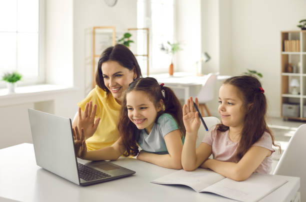 mamá y sus dos hijas se sientan frente a una computadora portátil y agitan una mano saludando a la maestra. - twin tips fotografías e imágenes de stock