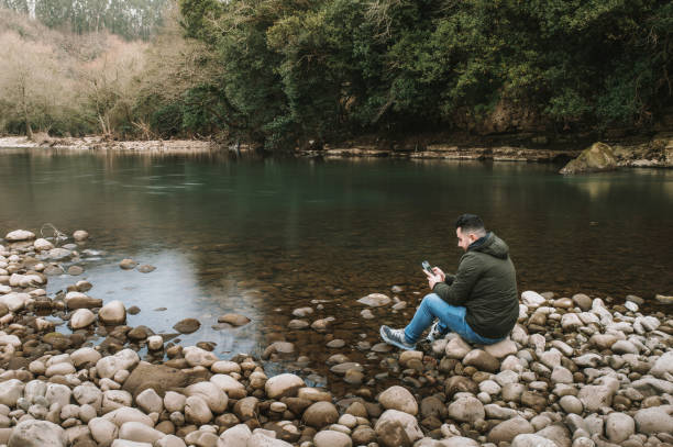 istock Young man sitting by a river using a smart phone 1367210900