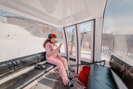 Woman skier using phone app in gondola ski lift on ski holidays. Girl smiling using mobile smartphone wearing cool ski clothing, helmet and goggles. Ski winter vacation activity concept