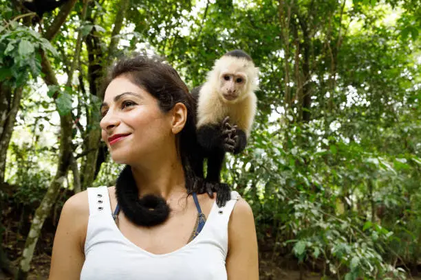 A capuchin monkey sits on a woman"u2019s shoulder.  The woman is smiling and looking to the left, the monkey is looking directly at the camera.  The woman has dark hair, is Iranian ethnicity, and looks happy.  The monkey has a combination of light and dark fur, it looks like it"u2019s probably enjoying itself too.  Photographed on a river tour in Costa Rica.