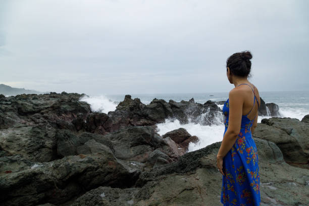 A woman in a blue dress walks on a rocky coastline.  The weather is overcast and dark.  The rocks are wet and waves crash against them.  She has dark hair pulled up in a bun, she is looking away from camera and out to sea.  The image has a melancholy mood.