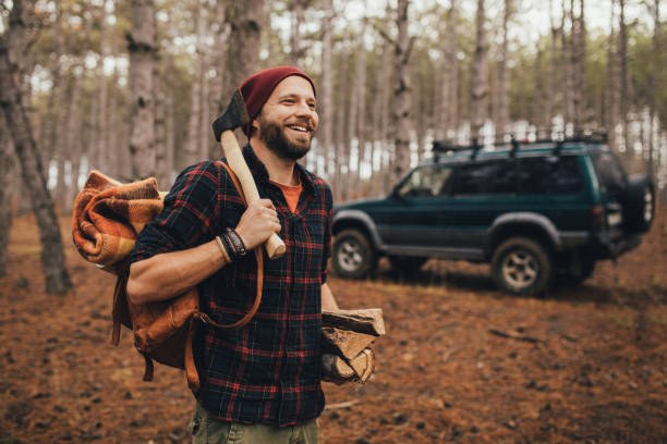 istock Millennial man hipster with beard wearing backpack, holding axe and firewood in forest with car on background. 1367215624