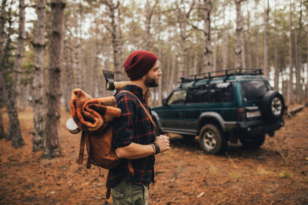 istock Millennial man hipster with beard wearing backpack, holding axe and firewood in forest with car on background. 1367215646