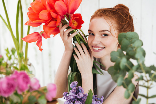 Young redhead woman florist looking up at camera and holding red tulips in workshop