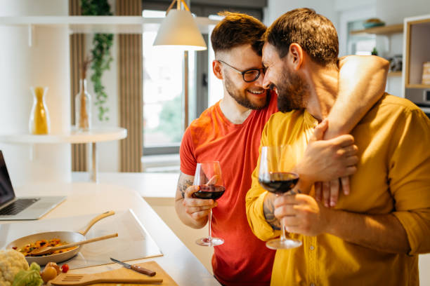 Happy gay couple in love hugging Happy hipster gay couple in love hugging while standing in the kitchen. They are cooking dinner together at home and drinking wine gay person stock pictures, royalty-free photos & images