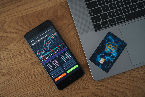 Smart phone with financial stock market analysis app and cryptocurrency credit card on a desk.