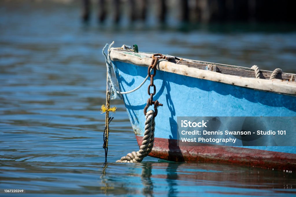 front of small blue row boat with anchor rope hanging off the front and disapearing in the calm watter Bow of small blue row boat anchored in the calm bay. White trim around the top. Blue base with a rust colored bottom which reflects in the calm water. Pier pilons out of focus in the background Anchor - Vessel Part Stock Photo