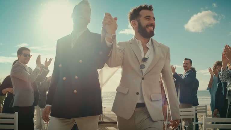 Handsome Gay Couple Walking Up the Aisle at Outdoors Wedding Ceremony Venue Near Ocean. Two Happy Men in Love Share Their Big Day with Diverse Multiethnic Friends. Cute LGBTQ Relationship Goals.