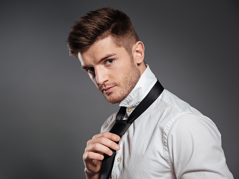 Picture of concentrated young man dressed in formalwear standing isolated over grey wall. Looking at camera.