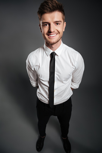 Top view full length portrait of a happy handsome man in formalwear standing and looking at camera isolated over grey background