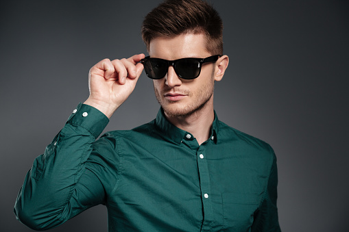 Picture of attractive serious man in sunglasses dressed in shirt posing over grey background. Looking aside.