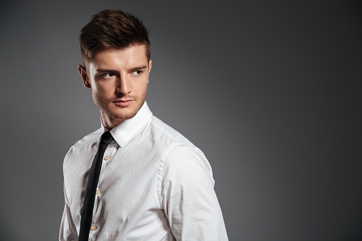 Serious handsome man in shirt and tie standing and looking away at copy space isolated over grey background