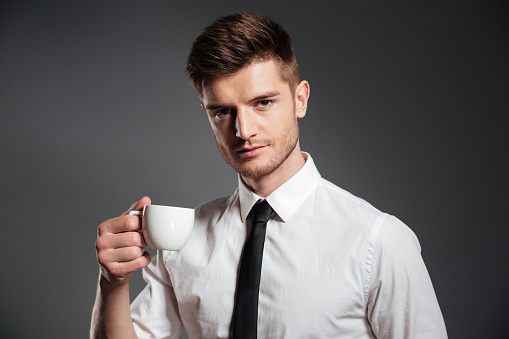 Close up portrait of a handsome young businessman with cup of coffee isolated over grey background