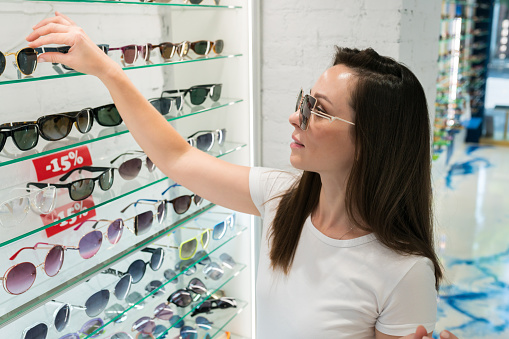 Thoughtful woman in white shirt looking at the display and choosing new sunglasses. Female customer shopping eyeglasses at local optical shop. Optics store concept