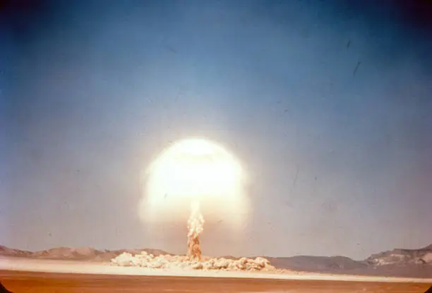 Vintage 35mm slide scan of a historic atom bomb explosion and mushroom cloud exploding in the middle of the desert. Mountain range on the horizon. Scratches on film. Dust cloud on the ground and tall mushroom cloud glowing and raining down