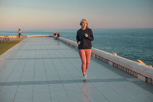 Asian sportswoman wearing black hoodie jogging on the embankment against the background of sea and listening to music in headphones