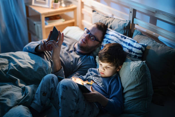 istock Little boy using digital tablet in bed at night while his father is using smartphone 1367240622