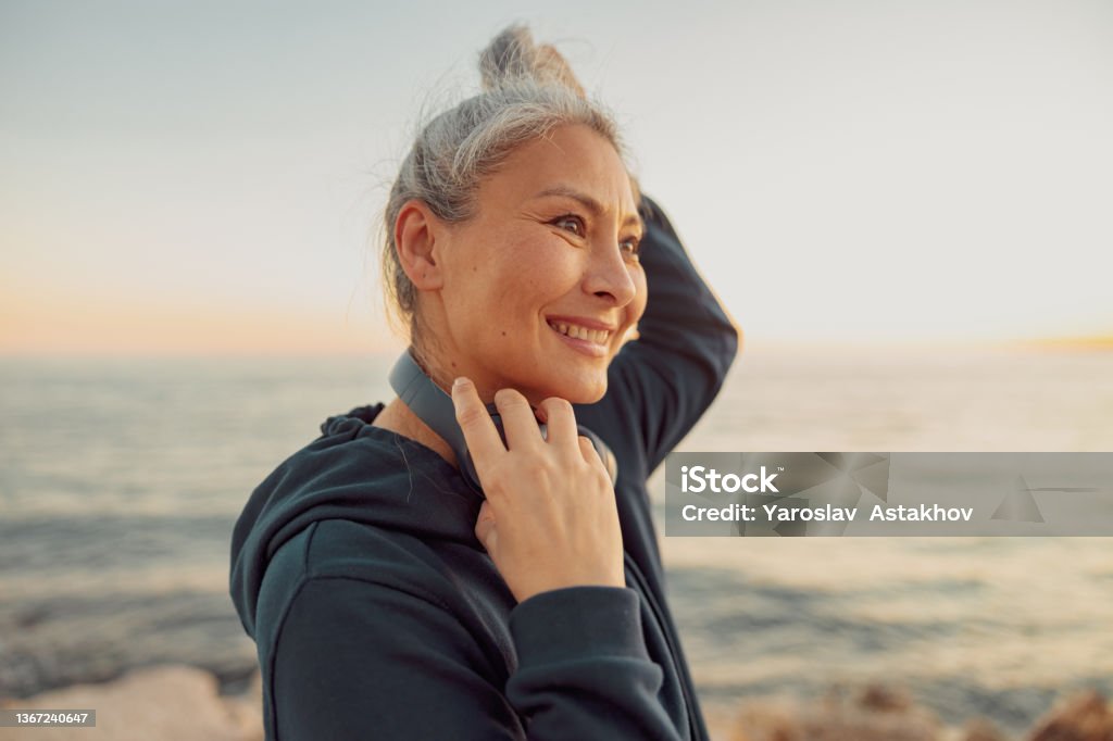 Charming sportswoman holding headphones, standing by the sea Close-up photo of mature Asian woman in black hoodie standing on the beach and wearing headphones, smiling One Woman Only Stock Photo
