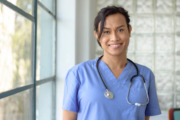 Female doctor of Asian descent A female doctor or nurse of Asian descent wearing scrubs and a stethoscope poses next to a window in a hospital corridor and smiles directly at the camera. self sacrifice stock pictures, royalty-free photos & images