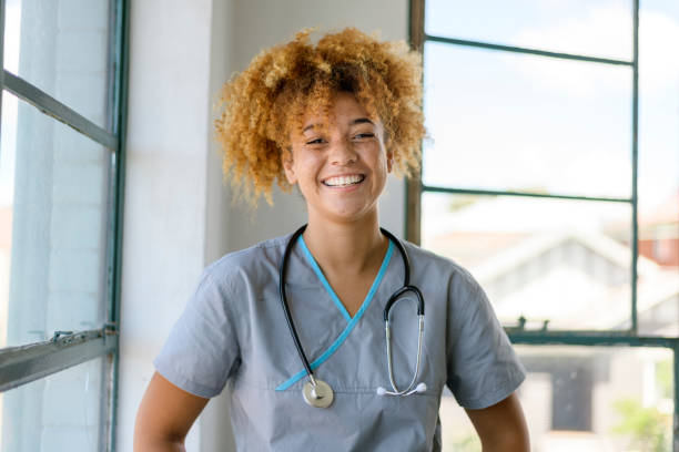 Friendly and young black female nurse A black female doctor or nurse wearing scrubs and a stethoscope poses next to a window in a hospital corridor and smiles confidently directly at the camera. self sacrifice stock pictures, royalty-free photos & images