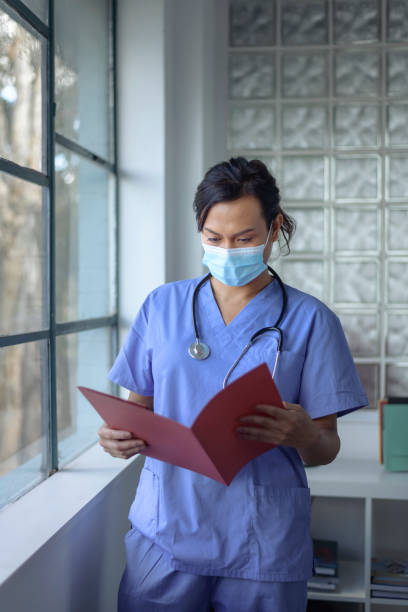 Female doctor reviewing patient medical records A female doctor of Asian descent wearing a protective face mask and scrubs looks at a file folder containing a patient's medical records before providing care while working in the hospital. self sacrifice stock pictures, royalty-free photos & images