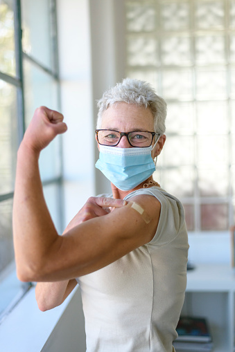 A senior adult woman flexes her arm and points at the bandage on her arm after receiving the COVID-19 vaccine injection at a medical clinic or hospital. The woman is wearing a protective face mask and is smiling directly at the camera.