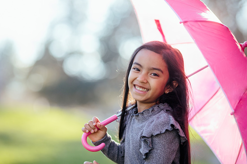 A smiling mixed ethnicity girl outside on a sunny day.  She holds a favorite pink umbrella.