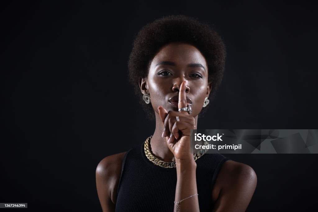 Closeup of beautiful young dark-skinned woman with finger on her lips showing shhh silence gesture on black background. Closeup of beautiful young african american dark-skinned woman with finger on her lips showing shhh silence gesture on black background. Women Stock Photo