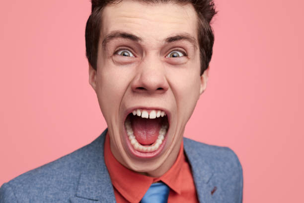 Enraged young businessman in suit looking at camera and shouting against pink background