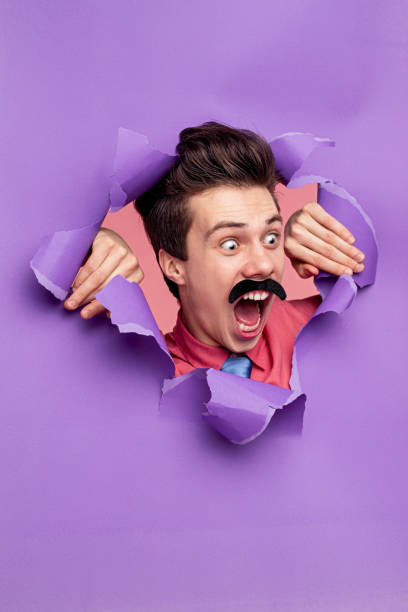 Funny man screaming through torn paper Crazy comic male with mustache shouting loudly while looking through ripped purple paper background in studio and looking away insanity stock pictures, royalty-free photos & images