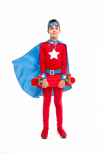 Full length strong boy in superhero costume carrying skateboard and looking at camera against white background