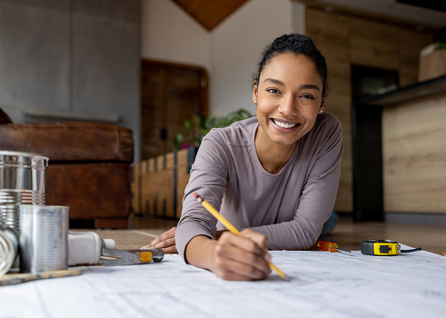 Happy African American female architect sketching some blueprints while working at home and looking at the camera smiling. **DESIGN ON BLUEPRINT WAS MADE FROM SCRATCH BY US**