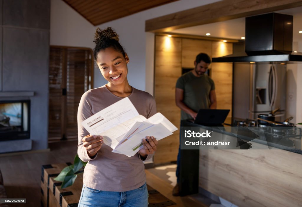 Woman at home looking at a utility bill that came in the mail Happy African American woman at home looking at a utility bill that came in the mail and smiling. **DESIGN ON DOCUMENTS WERE MADE FROM SCRATCH BY US** Financial Bill Stock Photo