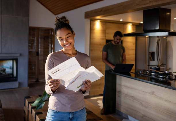 Woman at home looking at a utility bill that came in the mail Happy African American woman at home looking at a utility bill that came in the mail and smiling. **DESIGN ON DOCUMENTS WERE MADE FROM SCRATCH BY US** mail stock pictures, royalty-free photos & images