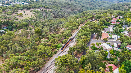 Aerial view luxury Overland Passenger Train departing for Melbourne, Sydney & Brisbane behind two bright orange locomotives passing through leafy hillside suburbs and natural bushland.  ID & logos edited. 16x9 format