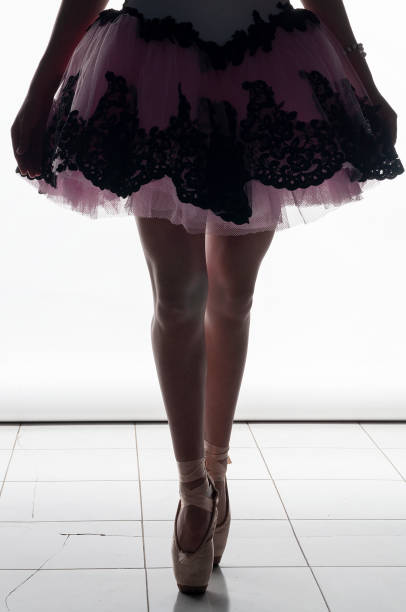 legs of a ballerina in a tutu, standing on tiptoe in her ballet shoes, in front of a white background and on top of a tile floor.
