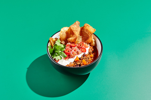 Chili con carne on colour background with hard shadow. Mexican chili in minimal style on green table. Traditional mexican cuisine on trendy design. Chili soup in bowl contemporary concept