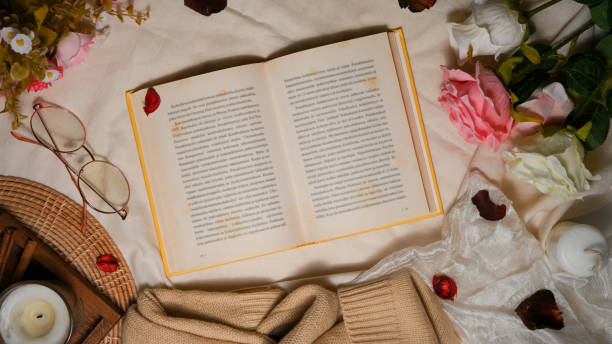 An open novel book in feminine vintage workspace. top view An open novel book in feminine vintage workspace with eye-glasses, flower bouquet, sweater, candles and decor on textile background. top view rose book stock pictures, royalty-free photos & images