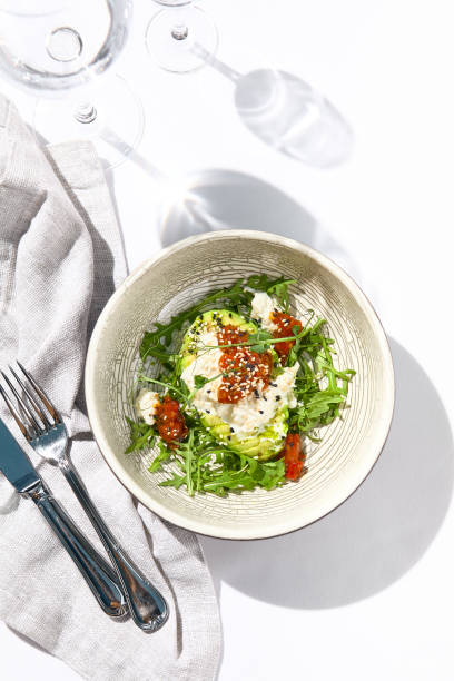 istock Avocado and stracciatella cheese salad. Fancy dinning with Italian salad bowl on white table with simple contemporary decor. Sunlight and harsh shadow still life. Avocado, cheese and tomato salad. 1367262336