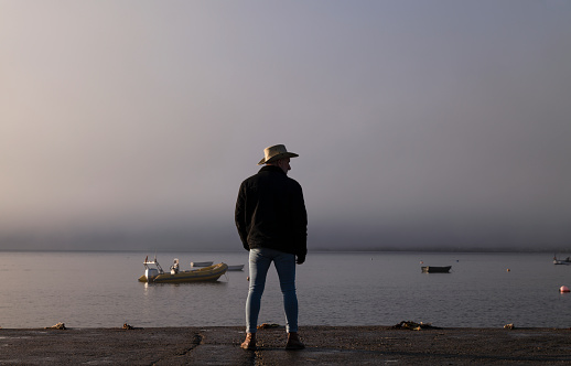 Portrait of adult man in cowboy hat and jeans standing on pier with fishing boats on sea. Shot in La Isleta del Moro, a small fishing village in Cabo de Gata Nature Park, Almeria, Spain