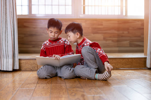 Asian brothers sitting on the floor in the living room and reading. They are wearing red sweaters.