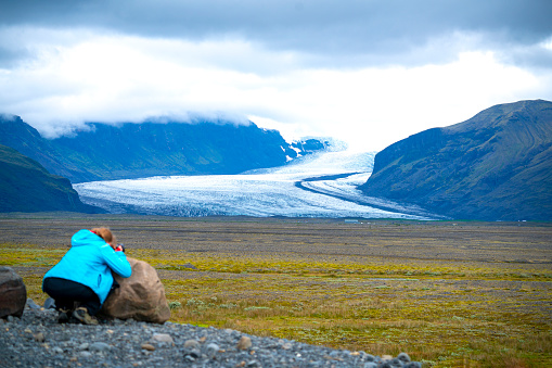 Senior woman kneels down on stone near circular road and photographing end of one line of Vatnajokull glacier in Skaftafell National park in Iceland. Upper part of photo is covered by clouds.plugs and photographs\nkneel down