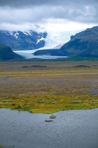 View from circular road of one line of Vatnajokull glacier in Skaftafell National park in Iceland. Upper part of photo is covered by clouds, front part small river water and in between is green grass.