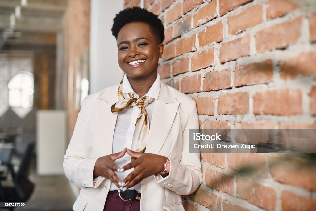 Confident black businesswoman leaning on a wall in the office and looking at camera. Satisfied African American businesswoman standing at corporate office and looking at camera. Businesswoman Stock Photo