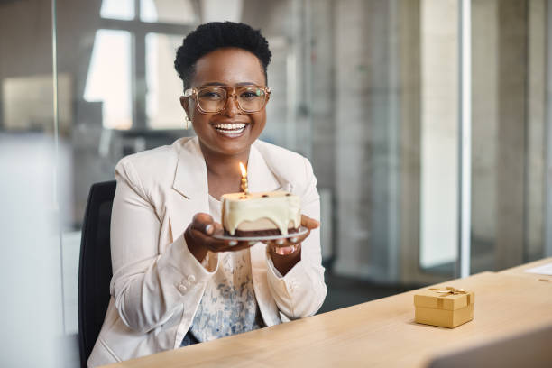 Happy African American businesswoman with her Birthday cake in the office looking at camera. Happy African American businesswoman holding cake with lightened candle in the office and looking at camera. office parties stock pictures, royalty-free photos & images
