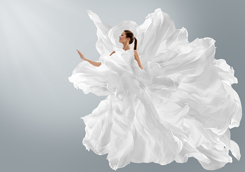 Modelo de moda en creativo vestido blanco puro como nube. Mujer con vestido largo de seda con tela de gasa volando sobre viento sobre fondo gris claro. Art Fantasy bailando Chica photo